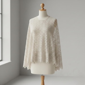 Puede incluir: Top de encaje color crema con cuello redondo y mangas largas y fluidas. El intrincado patrón de encaje floral cubre toda la prenda. El top se muestra en un maniquí, mostrando su delicada textura y diseño. Una prenda ligera y aireada.