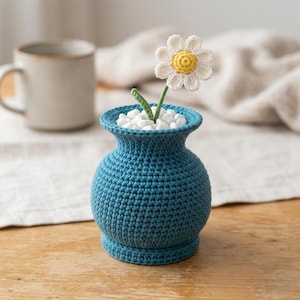 May include: A handcrafted, teal blue crochet vase holds a white and yellow daisy. The vase is filled with white pebbles. A beige mug sits in the background on a wooden surface. The crochet work is visible and detailed.