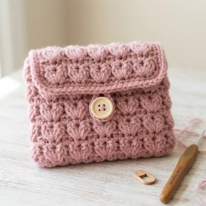 Könnte beinhalten: Handgefertigte, altrosa gehäkelte Tasche mit herzförmigem Muster. Die Tasche hat einen Klappenverschluss, der mit einem hellen Holzknopf gesichert ist. Ein Häkelhaken und ein Maschenmarkierer aus Holz sind sichtbar.