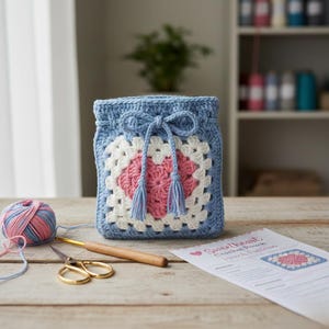 May include: A blue crocheted pouch with a white and pink heart design. The pouch has a drawstring closure with tassels. A ball of yarn, a crochet hook, and scissors are on the wooden surface. A pattern sheet is also visible.