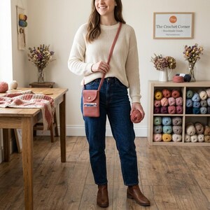 May include: A coral-colored crossbody phone bag with a flap and button closure. The bag is worn over the shoulder. The person is wearing a cream-colored sweater and blue jeans. A ball of yarn is held in the hand. The background includes a wooden table and shelves with yarn.