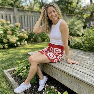 May include: A woman wearing a white tank top and red crochet shorts with a white geometric pattern. She is sitting on a wooden deck, wearing white sneakers, and smiling at the camera. The background includes greenery and a wooden fence.