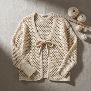 May include: A cream-coloured, open-weave crochet cardigan with long sleeves and a tie closure. The cardigan has a V-neck and ribbed cuffs. Crochet yarn and a hook are in the background, suggesting a handmade item.