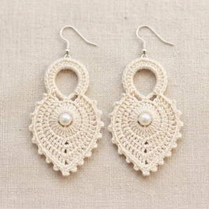 Puede incluir: Un par de pendientes de ganchillo de color crema. Cada pendiente presenta un diseño en forma de lágrima con una perla central y un borde festoneado. Los pendientes tienen ganchos para las orejas de color plateado.