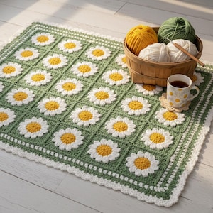 May include: A handmade crochet blanket featuring a daisy pattern in white, yellow, and green. The blanket has a scalloped white border and is displayed with yarn, a teacup, and a coaster.