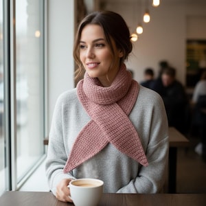 May include: A dusty rose-colored knitted scarf is draped around the neck. The scarf is made of a thick, textured yarn and has a pointed end. The person is wearing a gray sweater and holding a white coffee cup.