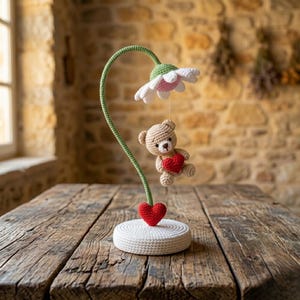 Puede incluir: Lámpara de ganchillo hecha a mano con un diseño caprichoso. La lámpara tiene un tallo verde, una flor blanca y rosa y un pequeño oso de peluche con un corazón rojo. La base es un diseño de ganchillo circular blanco. La lámpara está sobre una mesa de madera.