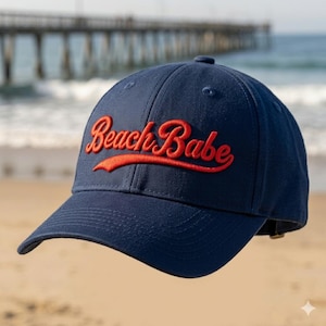 May include: A navy blue baseball cap featuring the text Beach Babe embroidered in a bold red script font across the front panels. The cap has a structured crown, a curved brim, and a button at the top center, showcasing a classic athletic design.