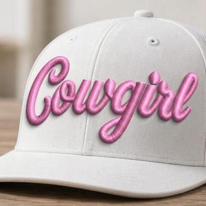 May include: White baseball cap with a curved brim and mesh back. The word "Cowgirl" is embroidered in a cursive, bubble-style font in a vibrant pink color across the front of the cap.