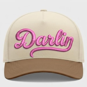 May include: A baseball cap with a cream-colored crown and a brown brim. The word "Darlin" is embroidered in a cursive, bubble-style font in a vibrant pink color across the front of the cap.