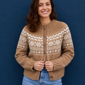 Könnte beinhalten: Eine beigefarbene Strickjacke mit weißem Fair-Isle-Muster an Brust und Schultern, rundem Halsausschnitt, langen Ärmeln mit gerippten Bündchen und einer Knopfleiste mit fünf braunen Knöpfen, getragen über einer hellblauen Jeanshose.