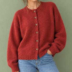 May include: A rust-colored, long-sleeved knit cardigan featuring a crew neckline and a front button-down closure with six small, round, light-brown buttons. The garment has a textured, slightly fuzzy knit finish and ribbed cuffs and hem.