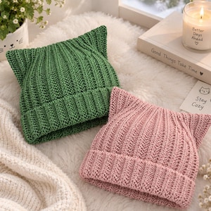 May include: Two knitted beanies with pointed cat-ear silhouettes, one in forest green and one in dusty pink. Both hats feature a vertical ribbed texture and a wide folded brim. The design creates a distinct feline shape at the top of each cap.