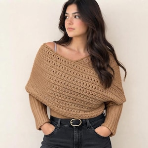 May include: A tan, long-sleeved, crocheted wrap top featuring a textured, horizontal stitch pattern with a crossover neckline design. The garment is worn over a light-colored base layer and paired with black jeans featuring a gold-toned metal belt buckle.