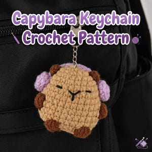 May include: A tan crochet capybara keychain with dark brown paws and ears, wearing light purple earmuffs. The capybara has embroidered black eyes and a nose. The text Capybara Keychain Crochet Pattern is displayed in white with a purple outline.