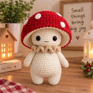 Puede incluir: Una muñeca de hongo tejida a ganchillo con un sombrero rojo grande y redondeado con puntos blancos, un cuerpo color crema y un cuello beige con volantes. La muñeca tiene dos pequeños ojos de cuentas negras y una boca cosida simple.