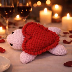 May include: A crocheted turtle with a red heart-shaped shell rests on a table set for a romantic dinner. Lit candles, wine glasses, and rose petals create a warm, intimate ambiance. Silverware is placed on the table.