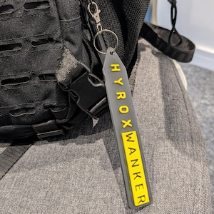 May include: A black tactical backpack with a gray and yellow keychain attached. The keychain reads "HYROXWANKER" in yellow letters on a gray background. The backpack features multiple compartments and straps.