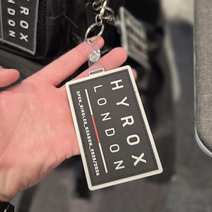 May include: A black and white HYROX LONDON tag with the text "OPEN_SINGLES_SEASON_2025/2026" is attached to a black bag. The tag is rectangular with a white border and a red stripe. The bag has additional HYROX patches.