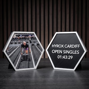 May include: Two hexagonal display plaques with white frames. The left plaque features a photograph of an athlete pushing a weighted sled on a track. The right plaque is black with white text reading HYROX CARDIFF OPEN SINGLES 01:43:29.