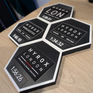 May include: A cluster of black and white hexagonal plaques, each featuring the word "HYROX" and times and locations including "LONDON" and "MANCHESTER". The plaques have white borders and display times such as 1:38:31, 1:46:10, 1:34:32, and 1:56:26.