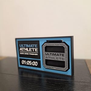 May include: A rectangular plaque with the words "ULTIMATE ATHLETE" and "SHOR 2000 FINISHER" in white text on a black background. The time "01:05:00" is displayed below. The plaque has a blue border and a silver-coloured centre with the words "ULTIMATE ATHLETE".