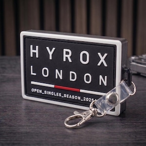 May include: A rectangular black and white badge featuring the white text HYROX LONDON and OPEN_SINGLES_SEASON_2025 with a thin red horizontal line. The badge has a clear plastic strap with a silver metal lobster clasp attached to the side.