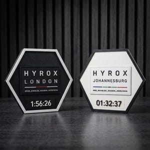 May include: Two hexagonal plaques with black and white frames. The left plaque is black with white text reading HYROX LONDON OPEN_SINGLES_SEASON_2025/2026 and 1:56:26. The right plaque is white with black text reading HYROX JOHANNESBURG PRO_DOUBLES_SEASON_2025/2026 and 01:32:37.