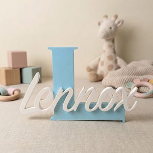 Puede incluir: Una letra "L" de madera azul claro con el nombre "Lennox" en escritura blanca. El nombre está adjunto a la letra. El fondo incluye una jirafa de peluche, bloques de madera y un juguete para bebés.