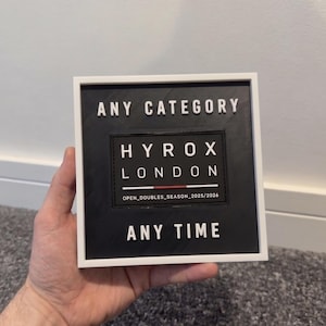 May include: A square black sign with white text that reads "ANY CATEGORY", "HYROX LONDON", and "ANY TIME". The sign also includes the text "OPEN_DOUBLES_SEASON_2025/2026". The sign is framed in white.