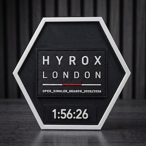 Peut inclure: Une plaque hexagonale noire avec une bordure blanche présentant le texte blanc HYROX LONDON, une ligne horizontale rouge et blanche, le texte OPEN_SINGLES_SEASON_2025/2026 et un affichage numérique en bas montrant les chiffres blancs 1:56:26.