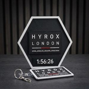 May include: A hexagonal black and white plaque featuring the text HYROX LONDON, a red line, and OPEN_SINGLES_SEASON_2025/2026, with the time 1:56:26 below. A matching rectangular keychain with the same text and design is placed in front.