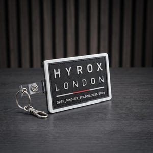 Può includere: Un portachiavi rettangolare bianco e nero con il testo HYROX LONDON in grassetto, una sottile linea orizzontale rossa e il testo OPEN_SINGLES_SEASON_2025/2026 in basso. L'etichetta è fissata a un anello metallico e a un moschettone.