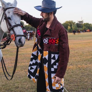 Op de afbeelding: Een donkerbruine suède jas met een lang silhouet, een reverskraag en een knoopsluiting. De jas is voorzien van panelen van doorgestikte stof met geometrische patronen in zwart, wit, geel en rood. Een donkergrijze cowboyhoed.