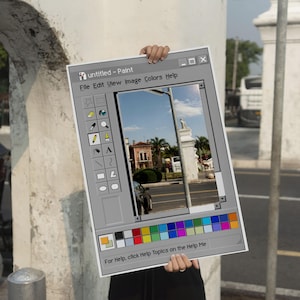 Op de afbeelding: Een persoon houdt een poster vast die lijkt op een klassiek Windows Paint-toepassingsvenster. De poster toont een straatbeeld met gebouwen en een blauwe lucht. Het Paint-venster bevat een werkbalk, een kleurenpalet en de tekst "untitled - Paint".