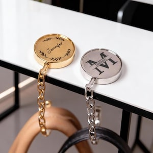 May include: Two personalized purse hangers. One is gold-toned with the name "Jennifer" and leaf accents. The other is silver-toned with the initial "M" and the name "Madison". Both have chain links and are designed to hang purses.