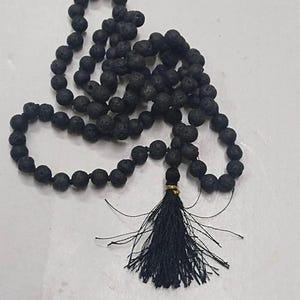 Puede incluir: Un collar largo compuesto por numerosas cuentas de lava negras, redondas y porosas, ensartadas juntas. El collar presenta una borla de hilo negro en la parte inferior, asegurada con un pequeño anillo de metal de color dorado brillante.