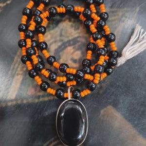 Pode incluir: Um colar de contas pretas e laranja. O colar tem um pingente grande, oval e preto com uma moldura de metal prateado. O colar é de várias voltas e tem uma borla.