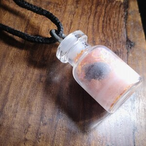 May include: A clear glass vial pendant filled with pink and black contents, suspended from a black cord. The vial has a clear stopper and is set against a wooden surface. The pendant is a unique accessory.