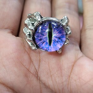 Puede incluir: Anillo plateado con un diseño de ojo grande, redondo, morado y azul. El ojo tiene una pupila negra vertical. El engaste del anillo tiene un diseño en forma de garra alrededor del ojo.