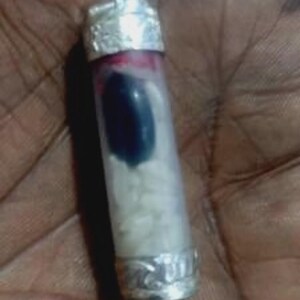 May include: A small, clear glass vial with silver-coloured metal caps at each end. The vial contains a dark blue object, white granules, and a red liquid. The vial is held in a hand.