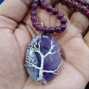 Può includere: Una collana con un ciondolo e perline viola. Il ciondolo è una pietra di ametista ovale con un design ad albero in filo d'argento. Le perline sono di un colore viola intenso e la collana è infilata su un cordino marrone.