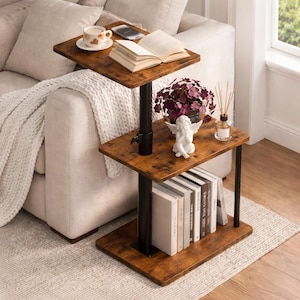 May include: A three-tiered side table with a distressed wood finish and black metal supports. The top tier holds a cup of coffee, a phone, and an open book. The middle tier has a small angel statue and a plant. The bottom tier holds books.