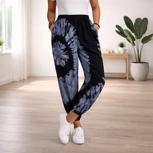 May include: Tie-dye trousers in black and grey with a spiral pattern on one leg and solid black on the other. The trousers have an elasticated waistband and pockets. The model is wearing white trainers.