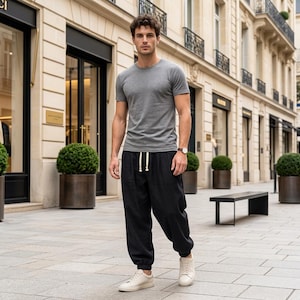 May include: A grey short-sleeved crew neck t-shirt paired with loose-fitting black trousers featuring an elasticized waistband with a white drawstring, and clean white low-top sneakers. A brown leather strap wristwatch is worn on the wrist.