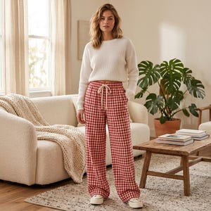 May include: A woman wearing a white waffle-knit sweater and red and white checkered wide-leg pants. The pants have a drawstring waist. She is wearing white sneakers. The background includes a couch, a plant, and a wooden coffee table.