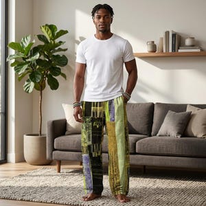 May include: A person wearing a white crew neck t-shirt and patterned pajama trousers. The pajama trousers feature a patchwork design with black, green, yellow, and purple vertical and horizontal stripes. The person is standing in front of a gray sofa and a large potted plant.