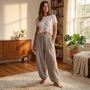 Könnte beinhalten: Eine Frau trägt ein weißes Oberteil mit Bindeband und eine hellbraune Haremshose mit Kordelzug und Knöchelbändern. Die Hose hat vertikale Streifen und Taschen. Das Outfit ist lässig und bequem.