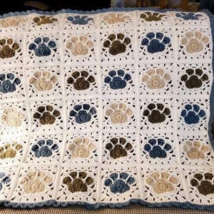 May include: A crocheted blanket featuring a repeating pattern of paw prints in shades of brown, beige, and blue on a white background. The blanket has a blue scalloped border, and is likely handmade.