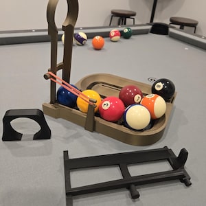 May include: A pool table setup with a variety of billiard balls and accessories. The balls are in various colors and numbered. A brown 3D-printed ball rack and a cue holder are visible. The table is gray with a green felt surface.
